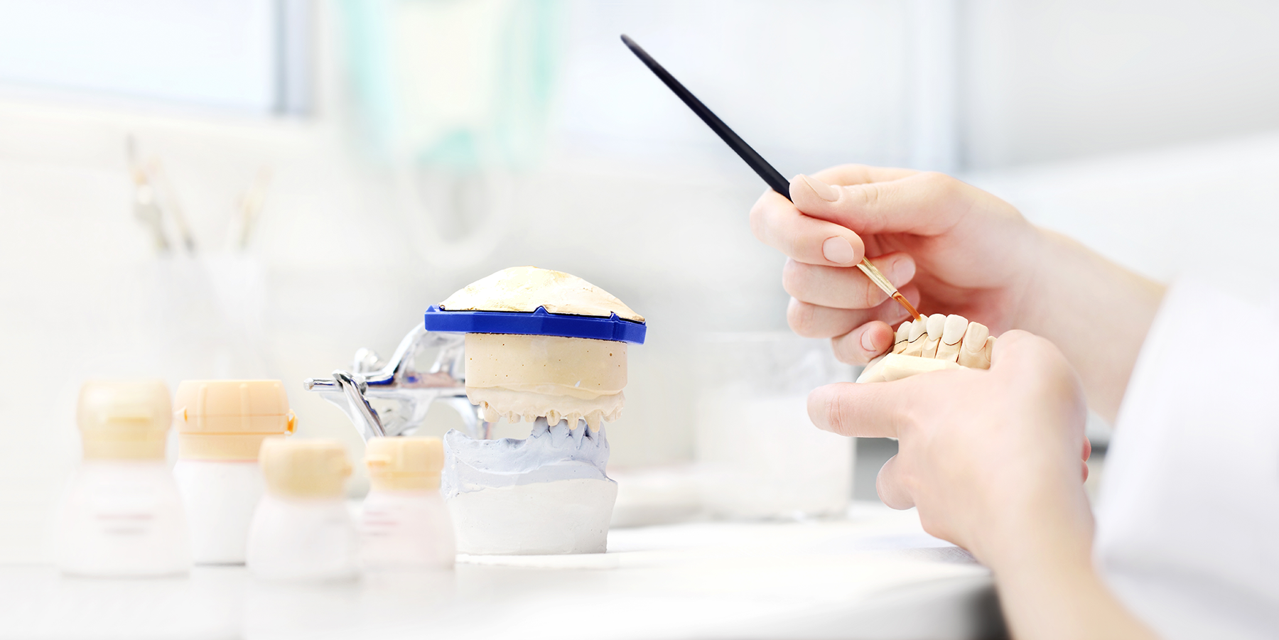 Dental technician's hands in lab working on denture model with paintbrush.